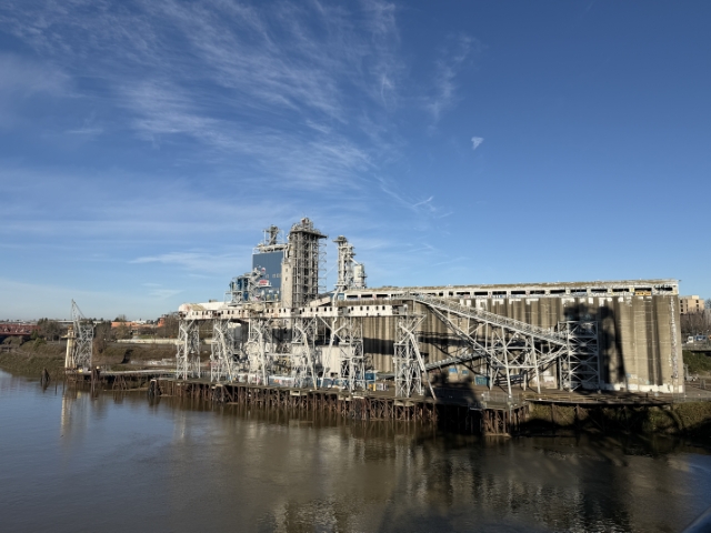 Industrial marine conveyor structures and docks at Portland's historic grain silos extending over the Willamette River, viewed from the west bank – December 2025.