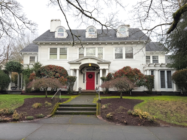 Exterior view of a well-maintained pre-1940 Colonial-style home in Portland, featuring a symmetrical facade, dormer windows, columns, red front door with wreath, and landscaped yard with steps leading to the entrance on an overcast day.