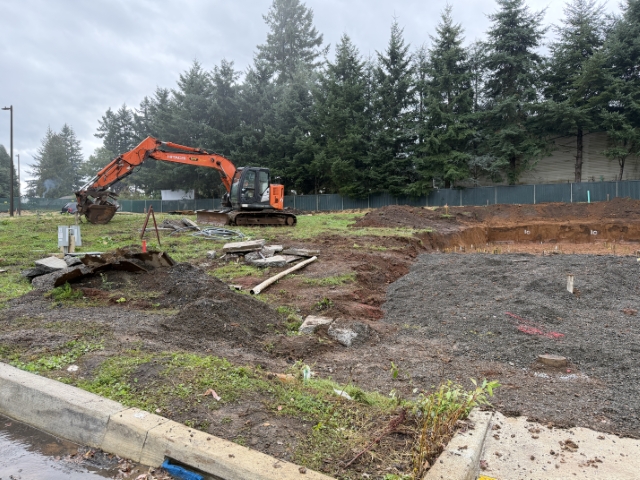 Active construction site in a vested pre-moratorium subdivision in Sandy, Oregon, showing an excavator digging on a muddy lot with foundation forms and dirt piles – illustrating ongoing development from allocated sewer connections during the moratorium.